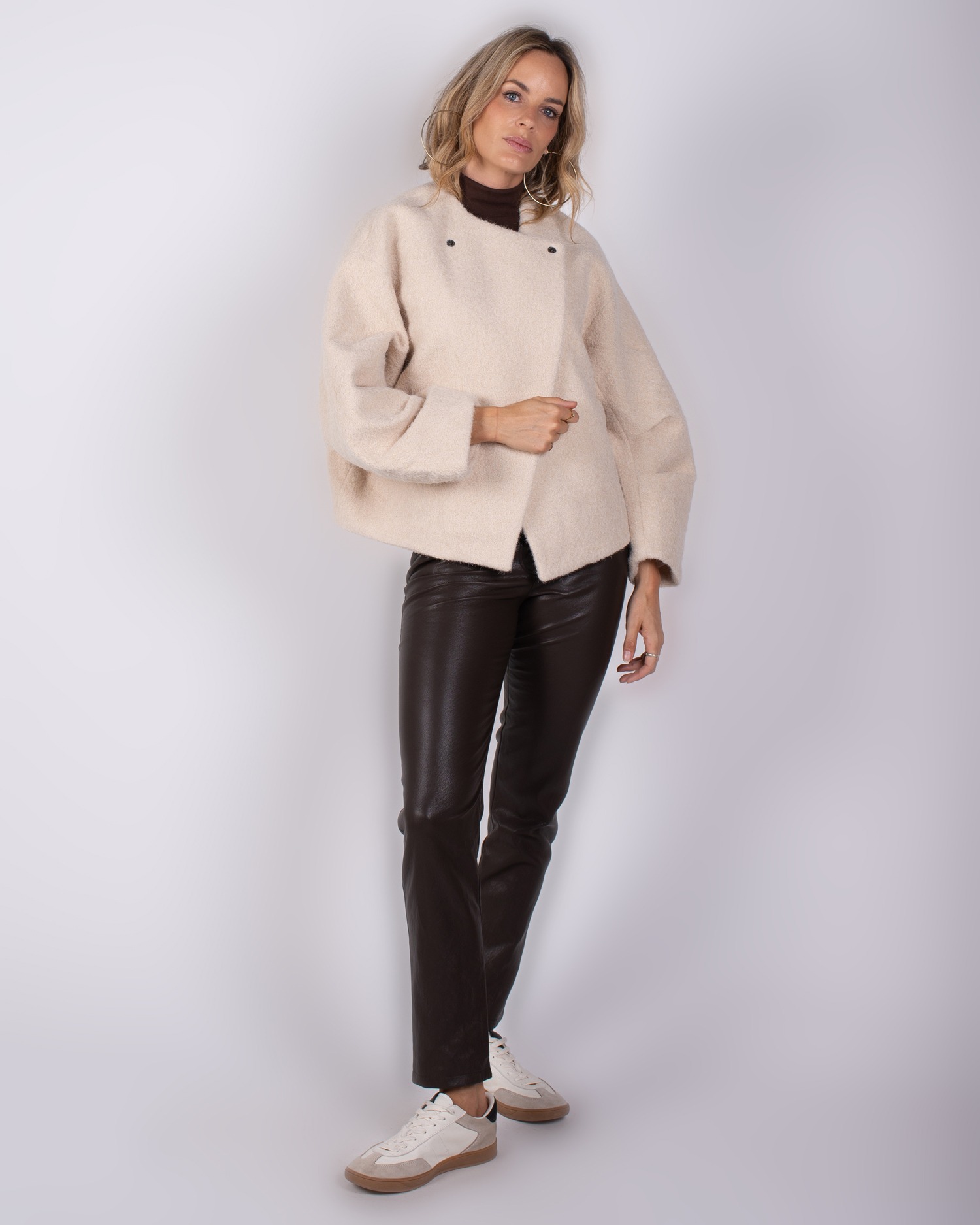 Studio_AW25-26#3-15 Vrouw draagt een beige oversized jas, donkerbruine leren broek en witte sneakers tegen een neutrale achtergrond.
