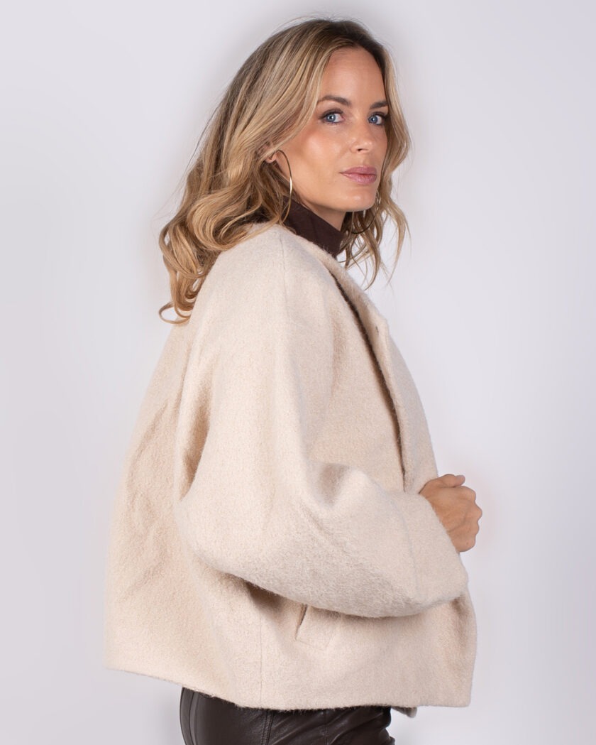 Vrouw in beige, wollen jas uit de Studio AW25-26 collectie, poserend in zijaanzicht tegen een lichte achtergrond.