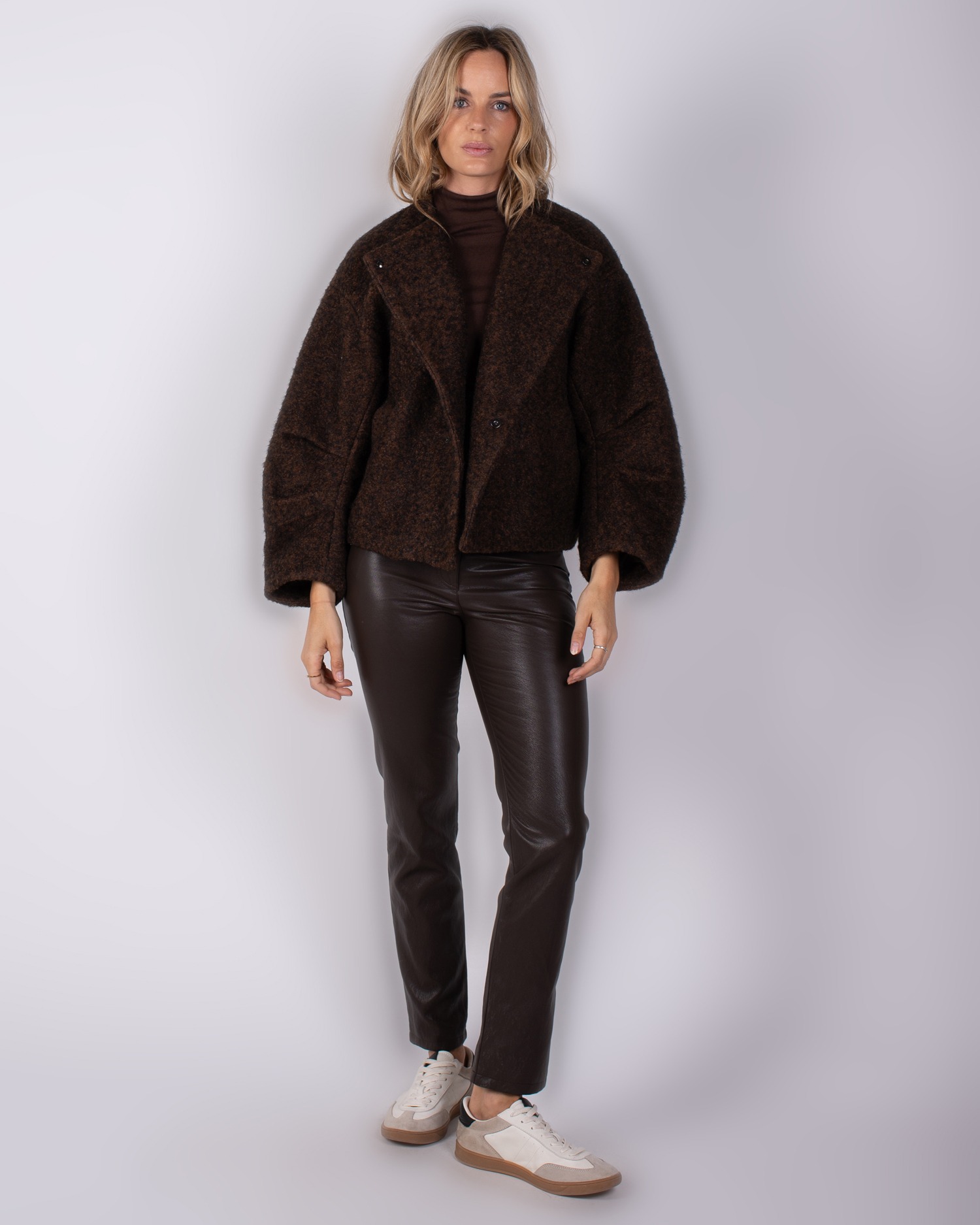 Studio_AW25-26#3-20 Vrouw in Studio_AW25-26#3-20 outfit met donkerbruine oversized jas, leren broek en witte sneakers.
