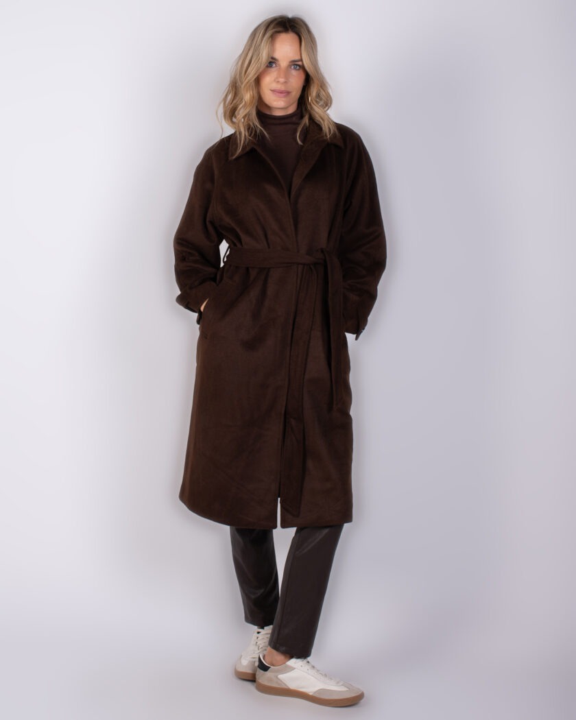 Vrouw draagt een donkerbruine, lange mantel met ceintuur uit de Studio AW25-26 collectie.