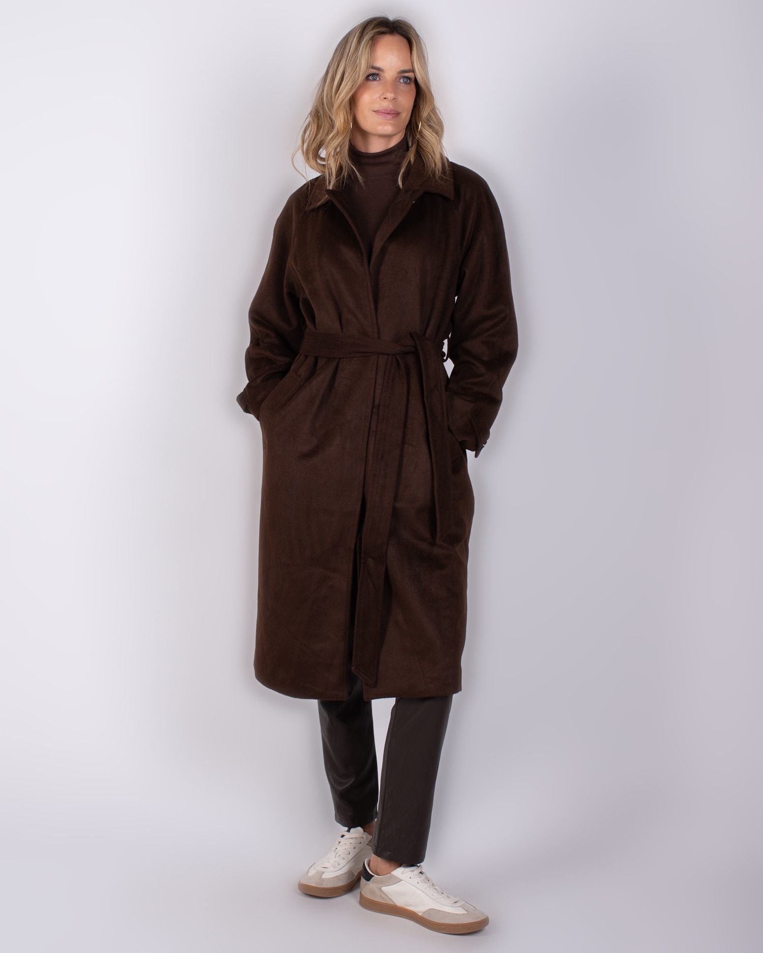 Vrouw draagt een lange, donkerbruine jas met ceintuur uit de Studio AW25-26 collectie.