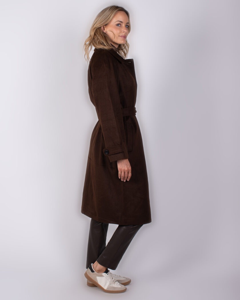 Vrouw in zijaanzicht draagt een donkerbruine, lange jas met ceintuur uit de Studio AW25-26 collectie.