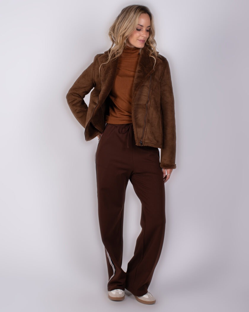 Vrouw draagt een bruine shearling jas met coltrui en donkere sportieve broek met witte streep.