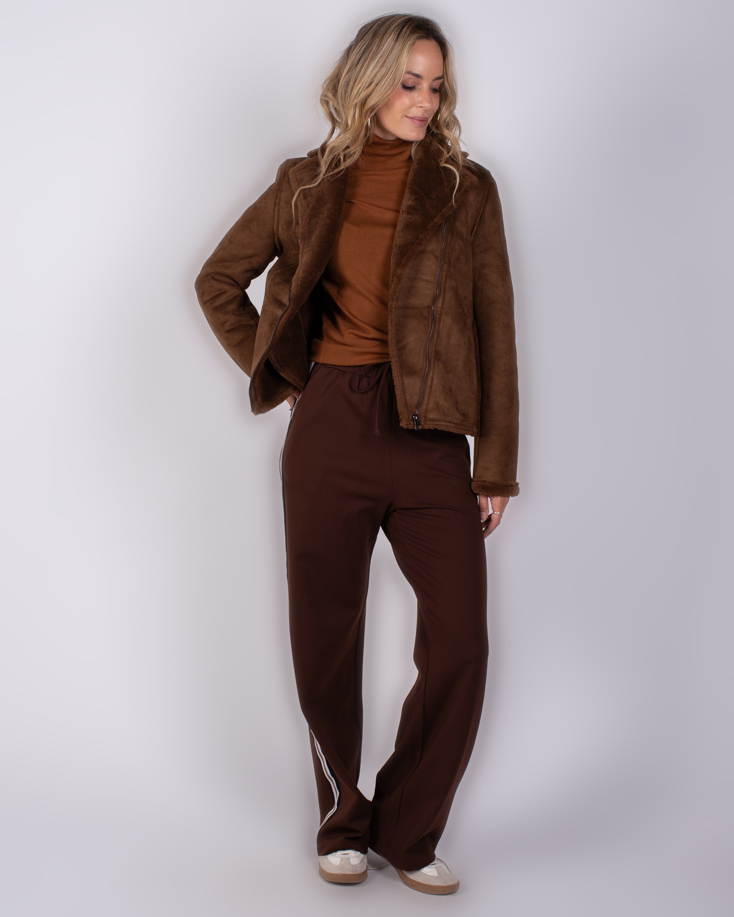 Vrouw draagt een bruine shearling jas met coltrui en donkere sportieve broek met witte streep.