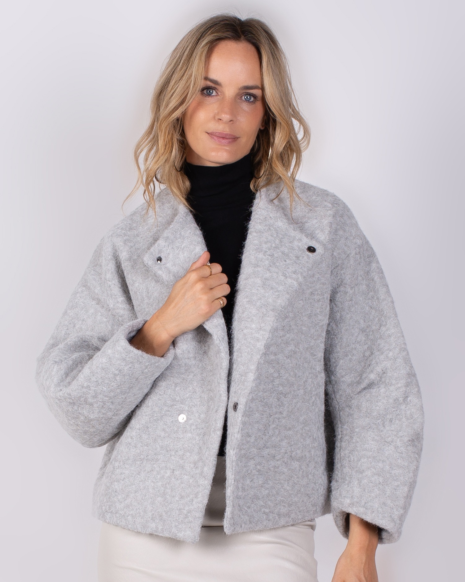 Vrouw met halflang blond haar draagt een lichtgrijze, wollige jas uit de Studio AW25-26 collectie.