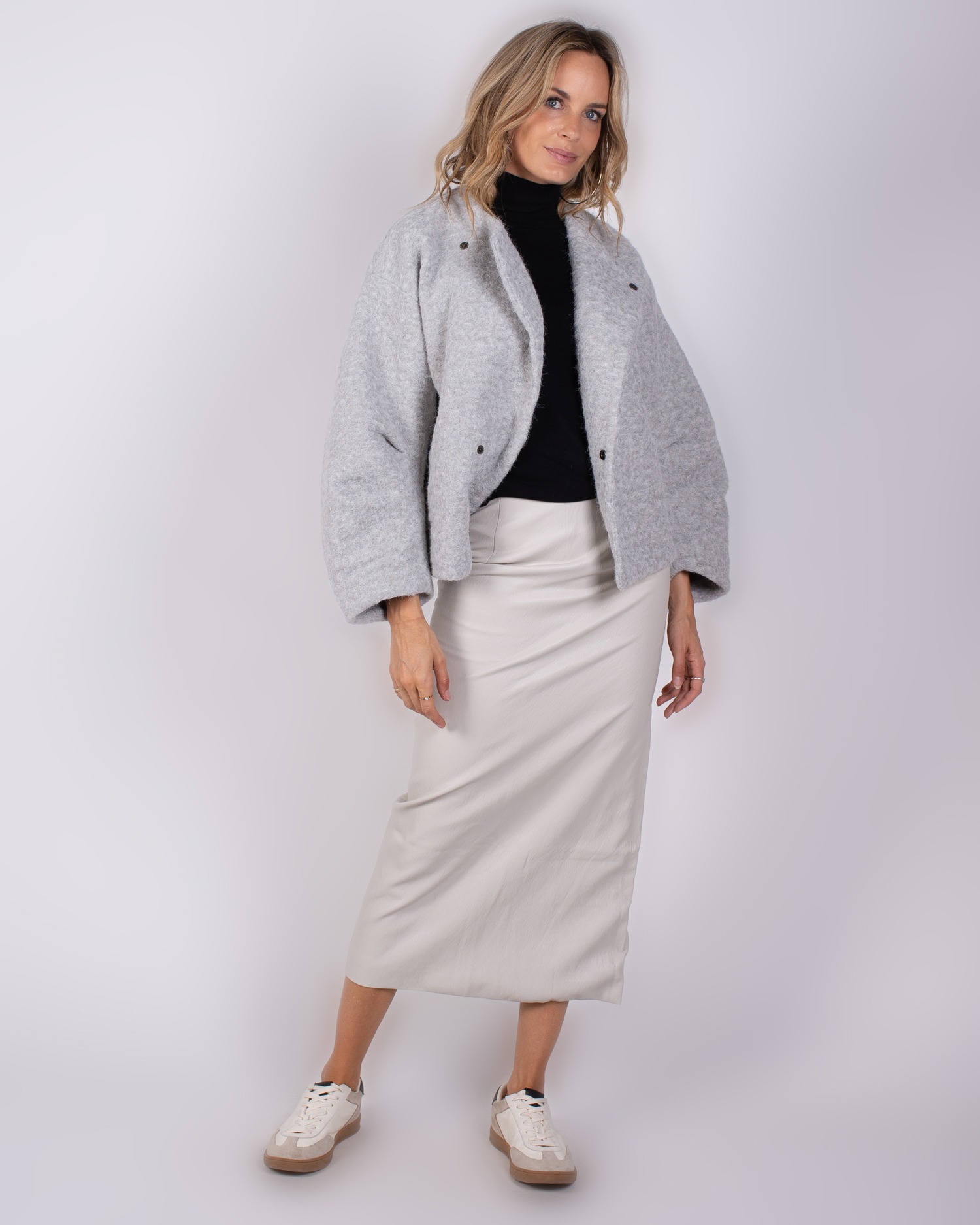 Studio_AW25-26#3-44 Vrouw draagt een lichtgrijze oversized jas, zwarte coltrui, crèmekleurige rok en sneakers uit de Studio AW25-26 collectie.