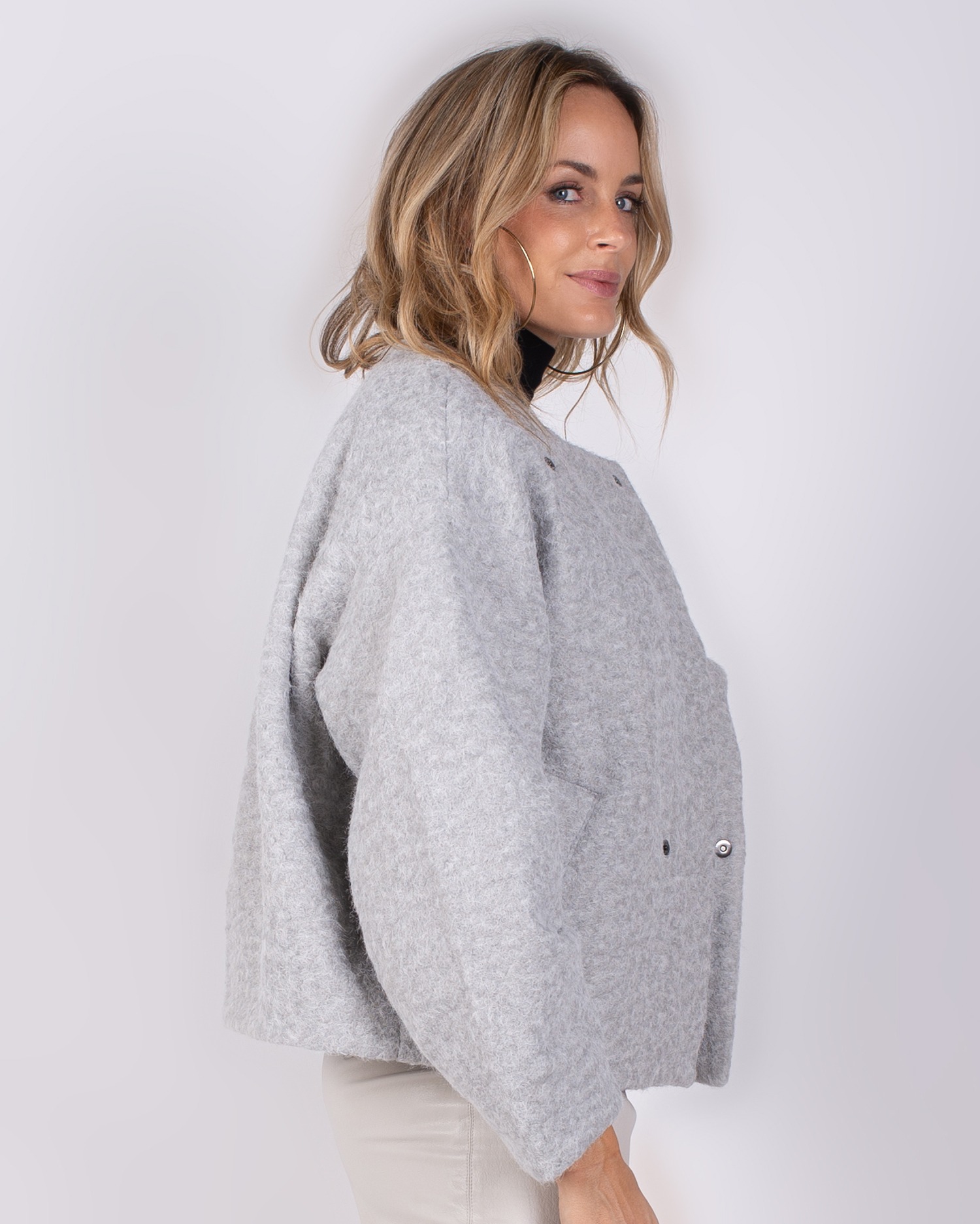 Studio_AW25-26#3-45 Vrouw poseert in zijaanzicht met een lichtgrijze, wollen jas uit de Studio AW25-26 collectie.