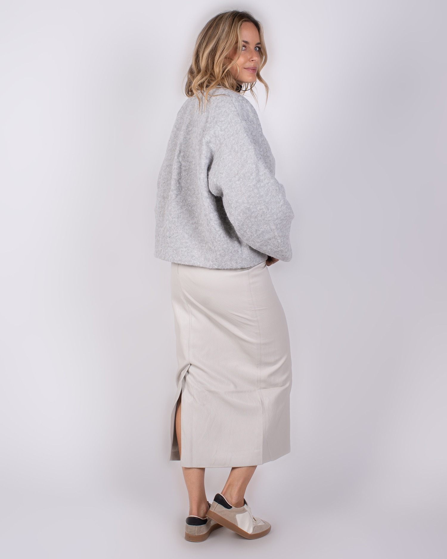 Studio_AW25-26#3-47 Vrouw in een lichtgrijze wollen trui en een beige rok met split, poserend zijwaarts voor een witte achtergrond.