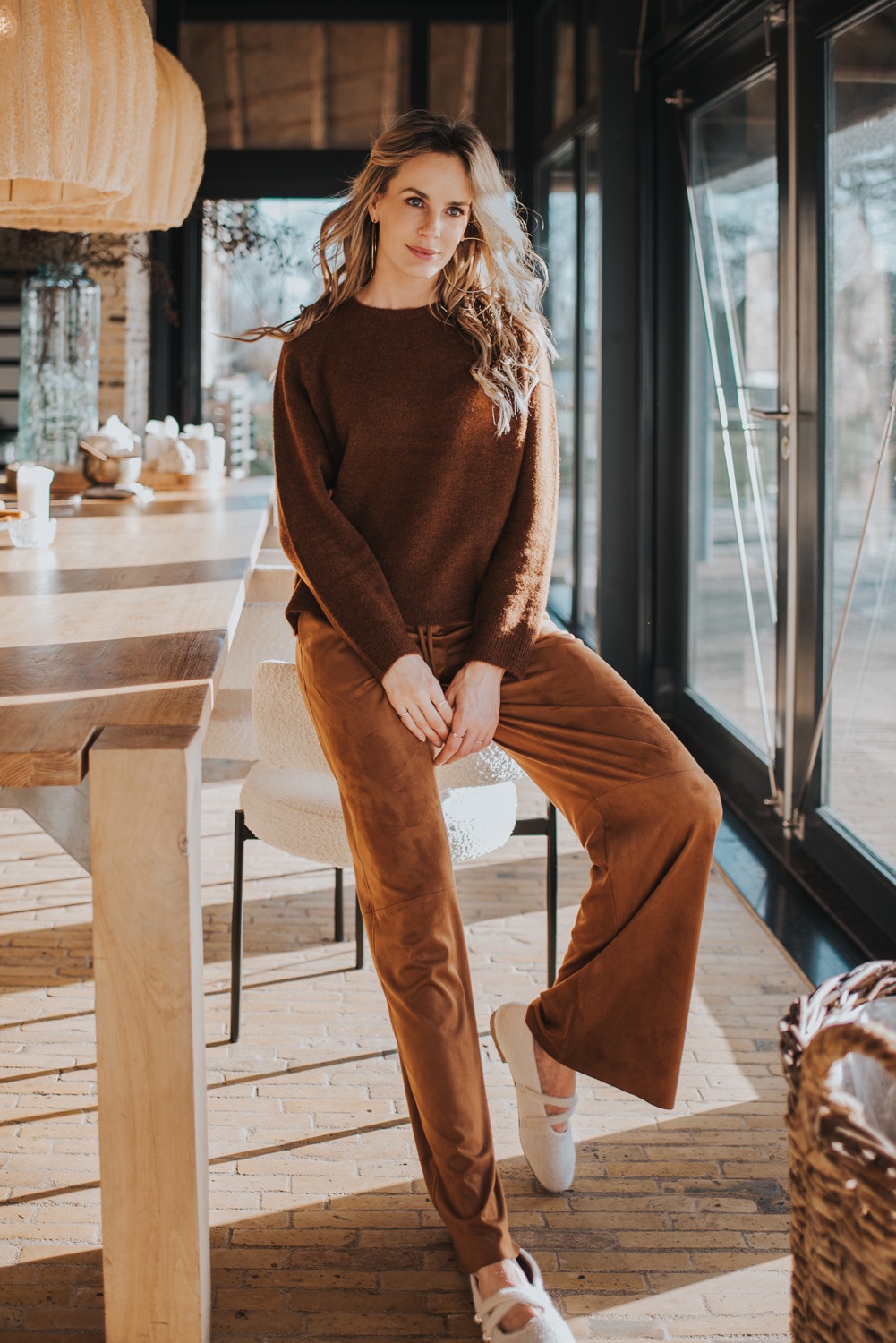 Vrouw poseert in een warme, bruinkleurige outfit uit de AW25-21 collectie in een sfeervolle, modern ingerichte ruimte.
