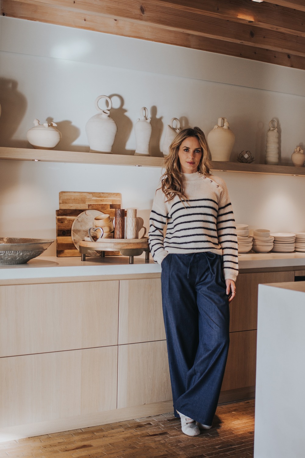Vrouw in een moderne keuken leunend tegen het aanrecht, gekleed in een gestreepte trui en wijde donkerblauwe broek (image_AW25-5).