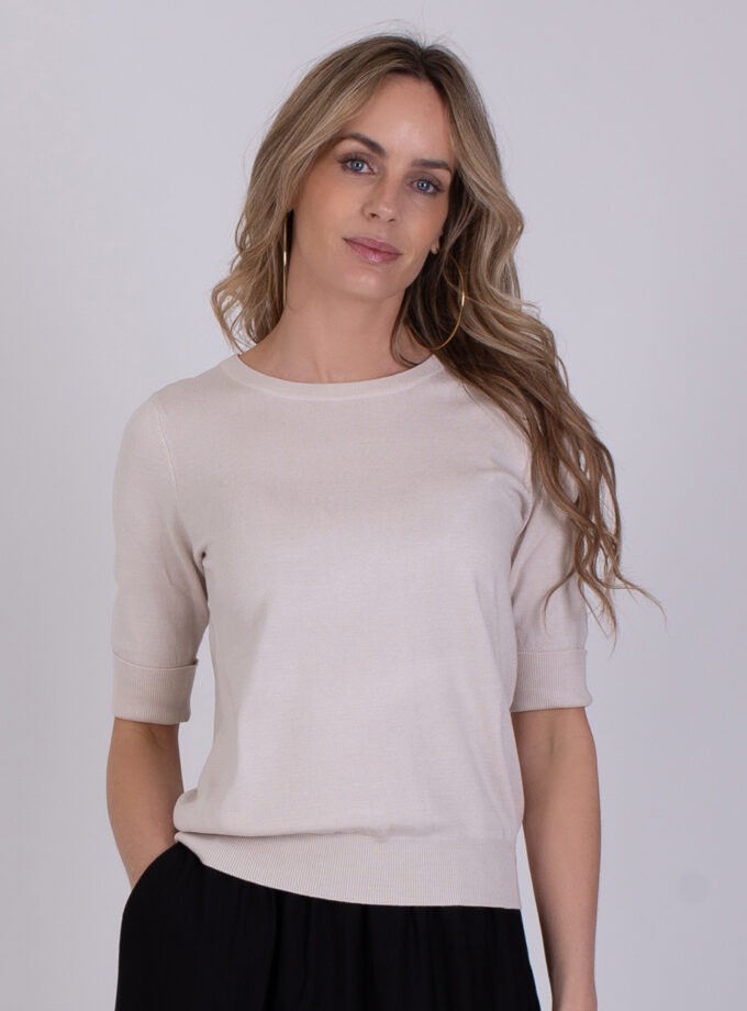 Vrouw draagt een licht beige Darwin Kit 2 trui met korte mouwen en een zwarte broek tegen een neutrale achtergrond.