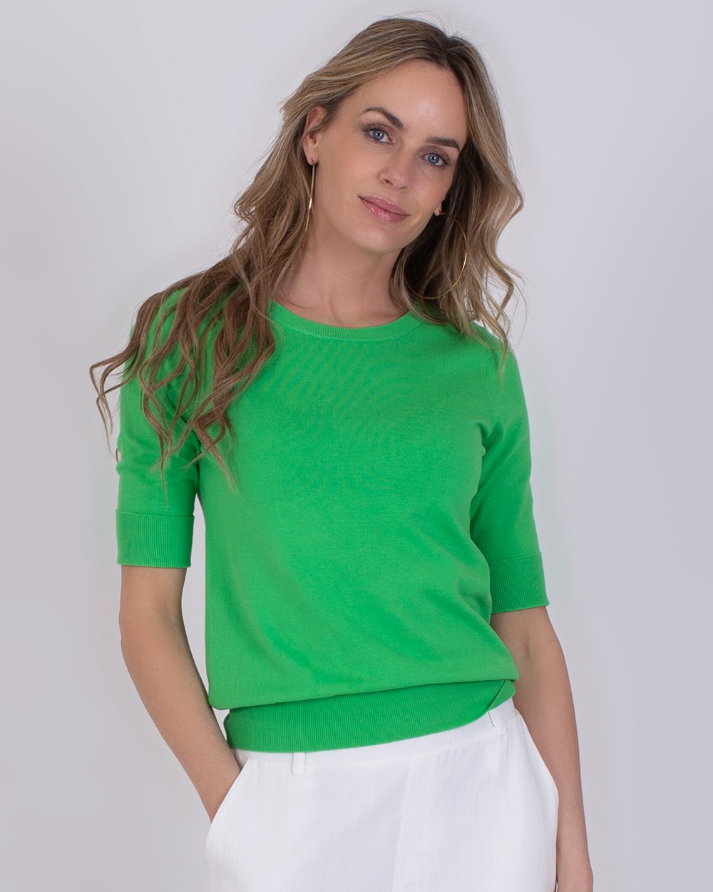 Vrouw draagt een groene Darwin Sea Green trui met halflange mouwen en witte broek.