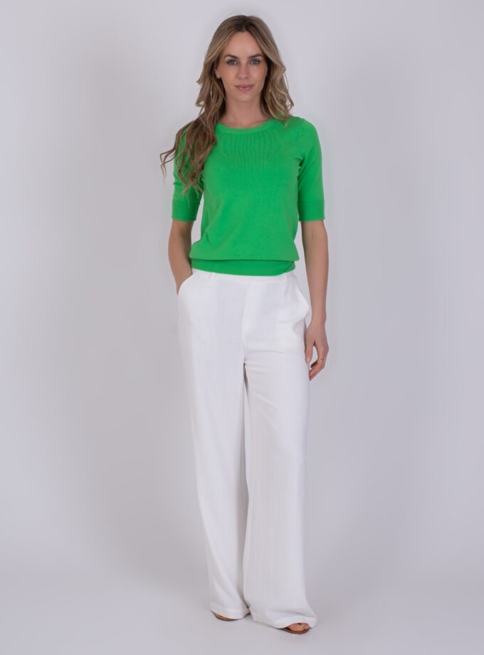 Vrouw draagt een felgroene Darwin Sea Green trui met halve mouwen en witte, wijde broek.