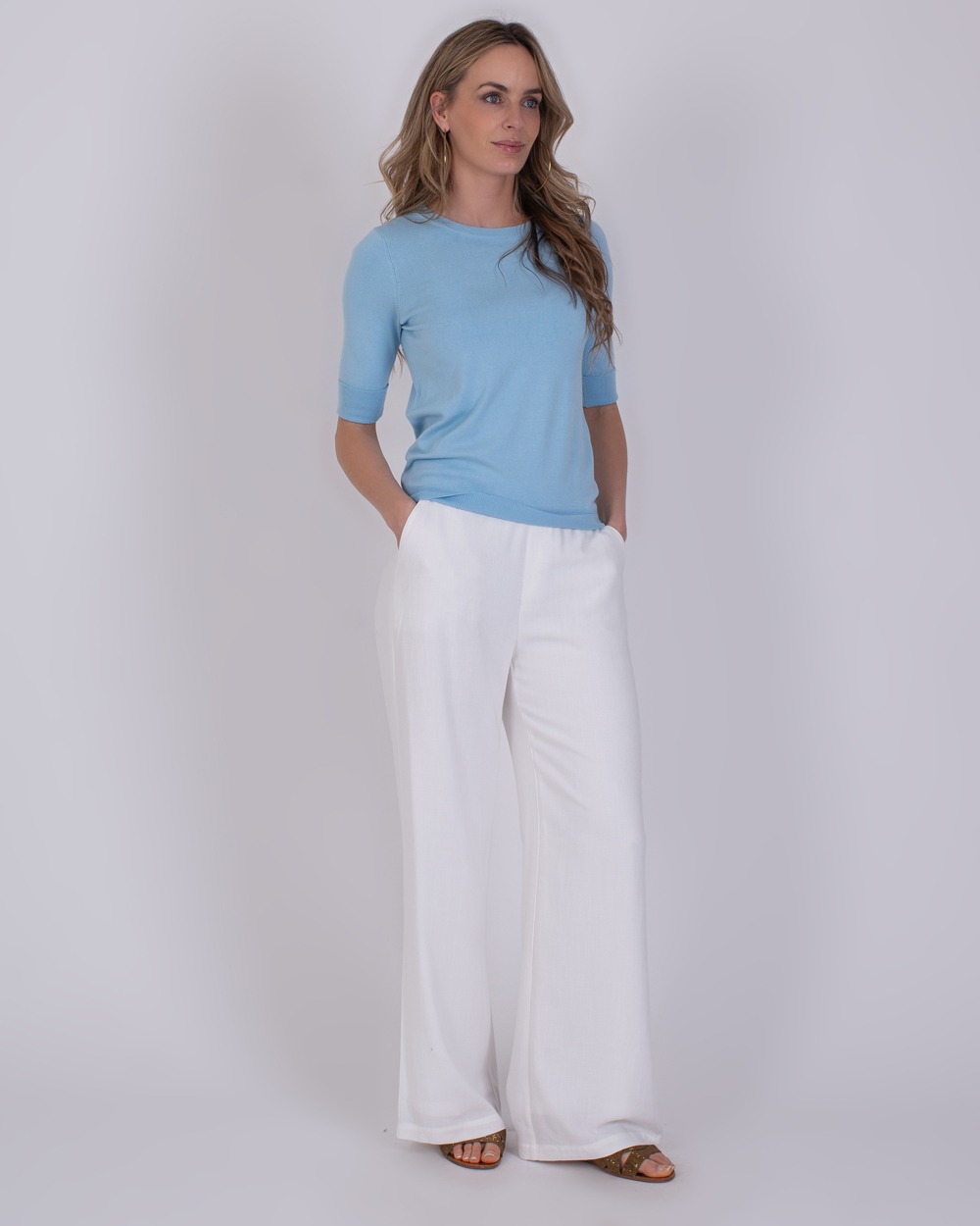 Darwin soft blue 2 Vrouw draagt een Darwin soft blue trui met half lange mouwen en een witte wijde broek.