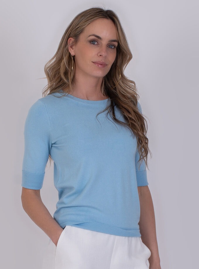 Vrouw draagt een lichtblauwe Darwin-trui met halve mouwen en een witte broek.