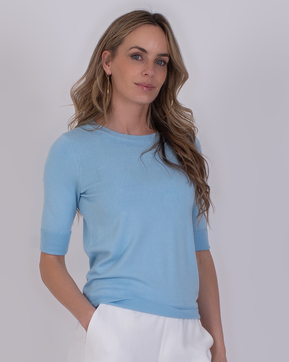 Vrouw draagt een lichtblauwe Darwin-trui met halve mouwen en een witte broek.