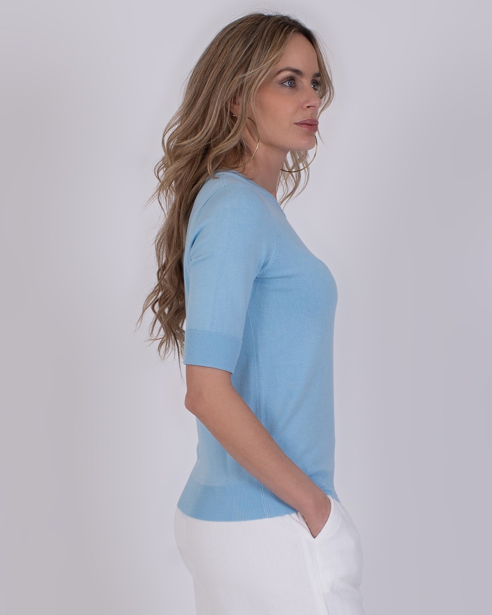 Darwin soft blue 4 Vrouw in zijaanzicht draagt een lichtblauwe Darwin soft blue trui met halve mouwen en witte broek.
