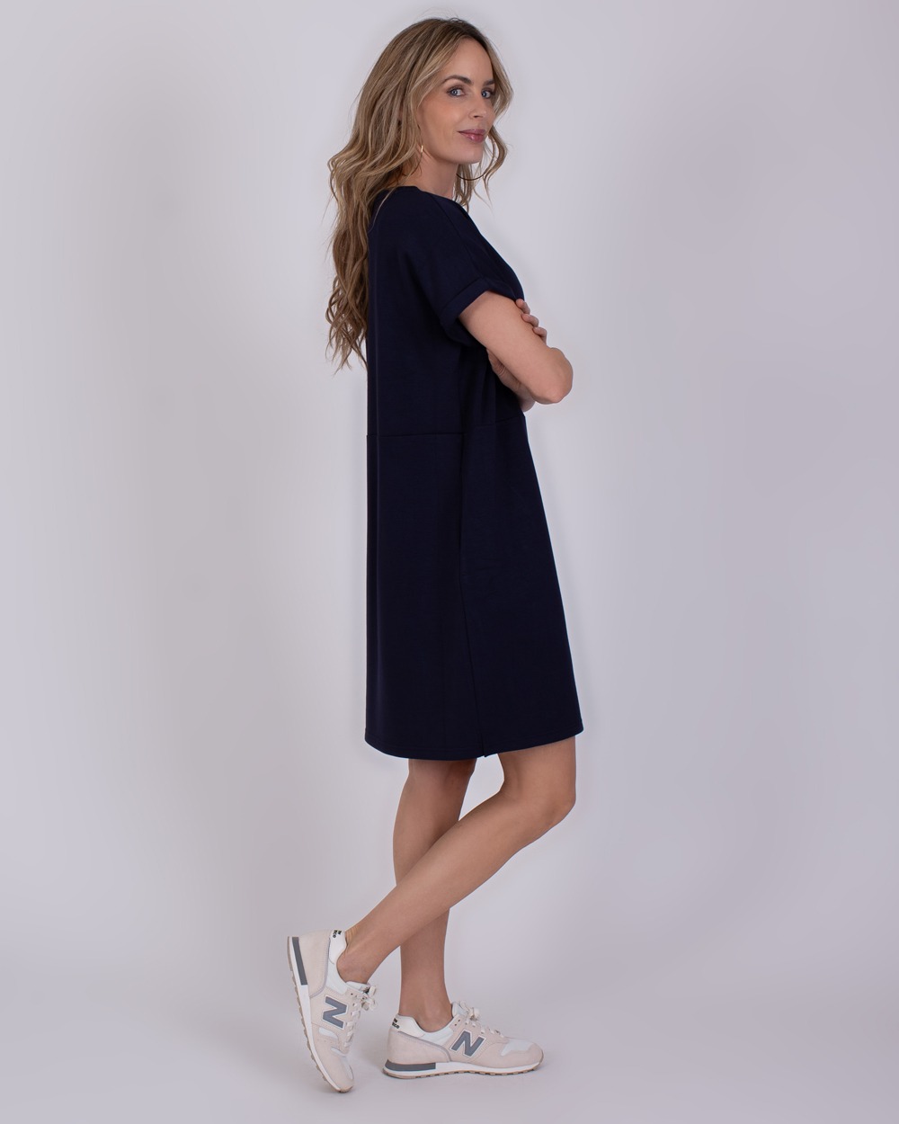 Liverpool navy 2 Vrouw in een donkerblauwe Liverpool-jurk en witte sneakers poseert zijwaarts tegen een neutrale achtergrond.