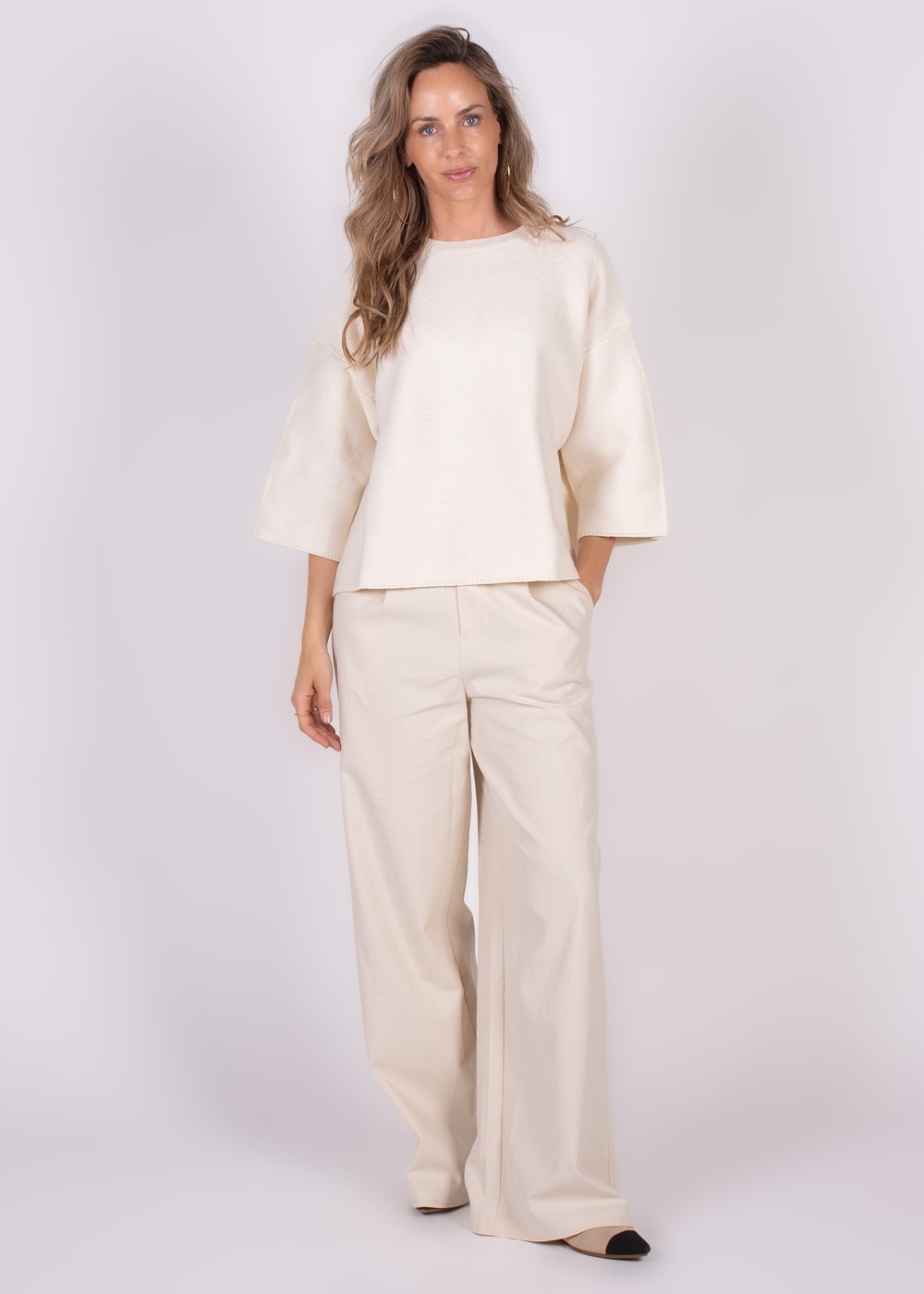 Een vrouw poseert in een offwhite outfit, bestaande uit een wijde trui en broek, tegen een neutrale achtergrond.