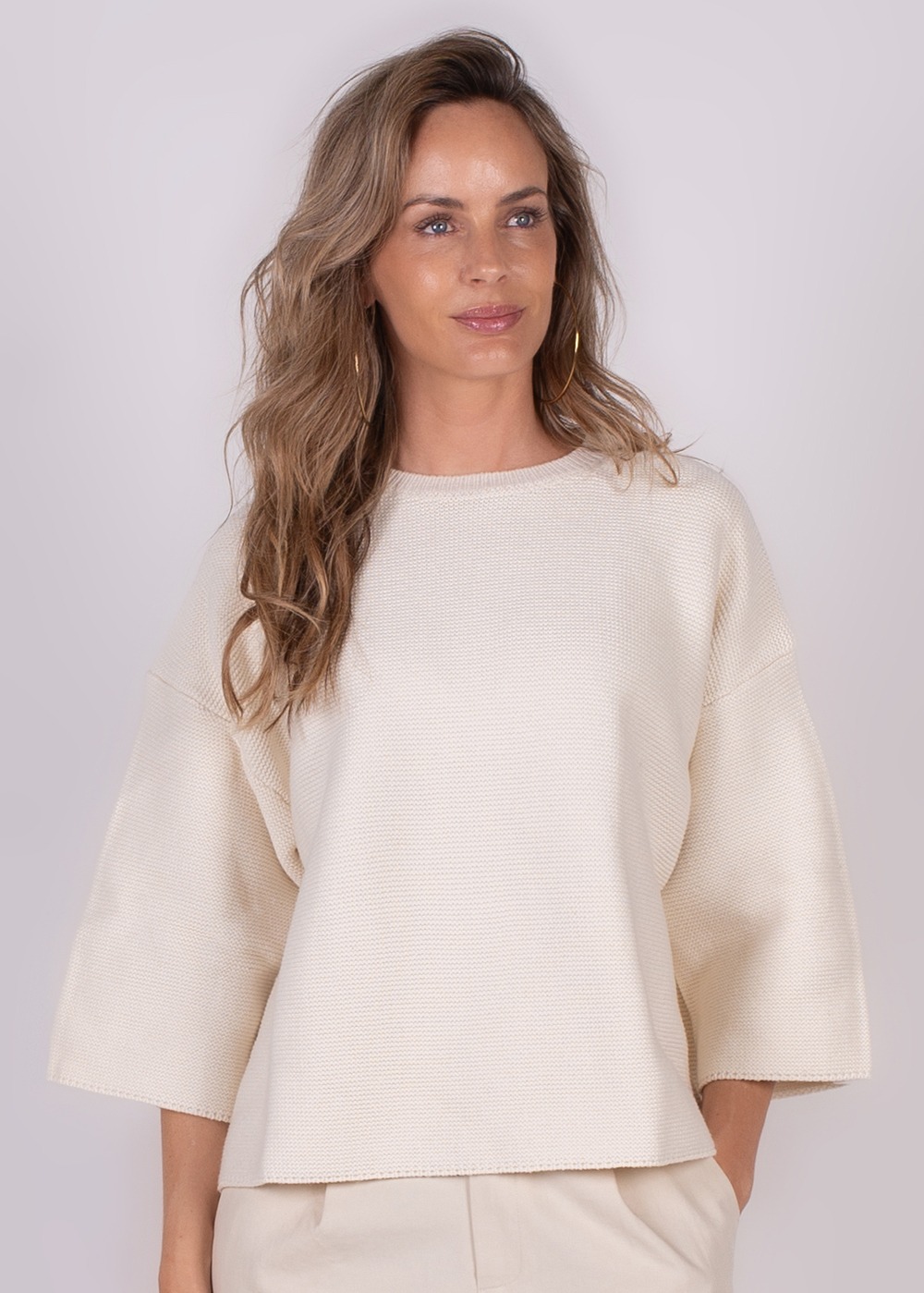 Een vrouw draagt een offwhite Wellington trui met wijde mouwen, tegen een neutrale achtergrond.