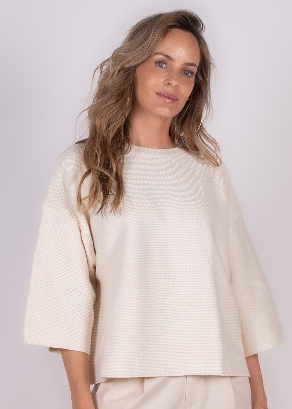 Een vrouw met lang bruin haar draagt een offwhite, ruime trui met driekwartsmouwen, tegen een neutrale achtergrond.