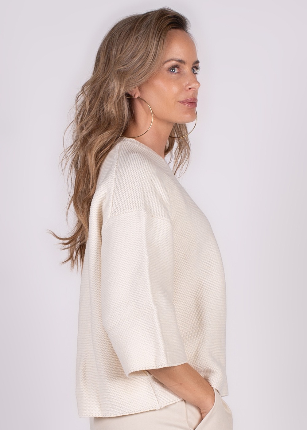 Een vrouw in profiel die een offwhite trui draagt, met grote oorringen en haar handen in haar zakken.