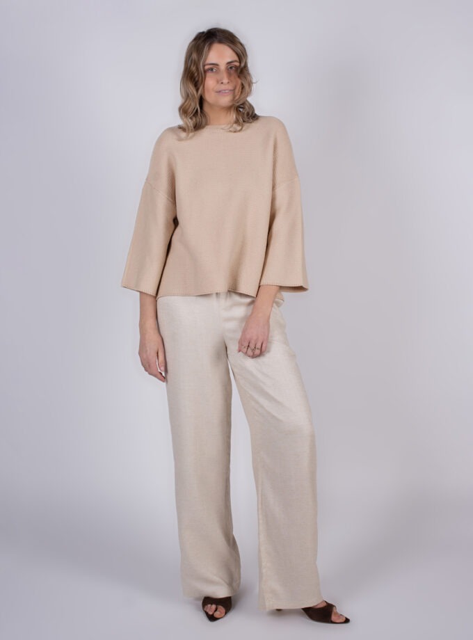 Model draagt een beige trui met wijde mouwen en een lichtgekleurde broek in een studio-omgeving.