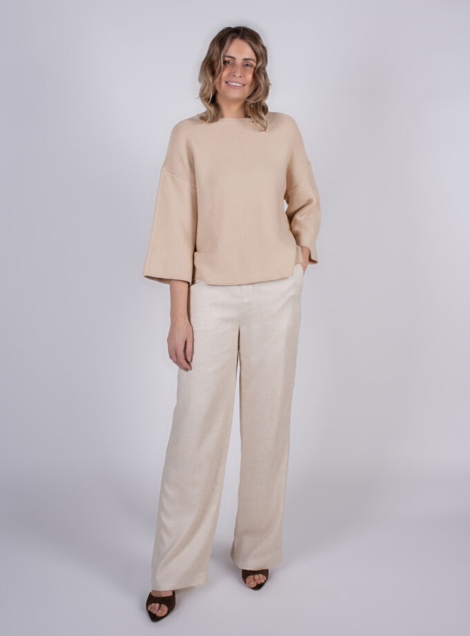 Vrouw poseert in een beige trui en wijde, lichte broek voor een neutrale achtergrond.