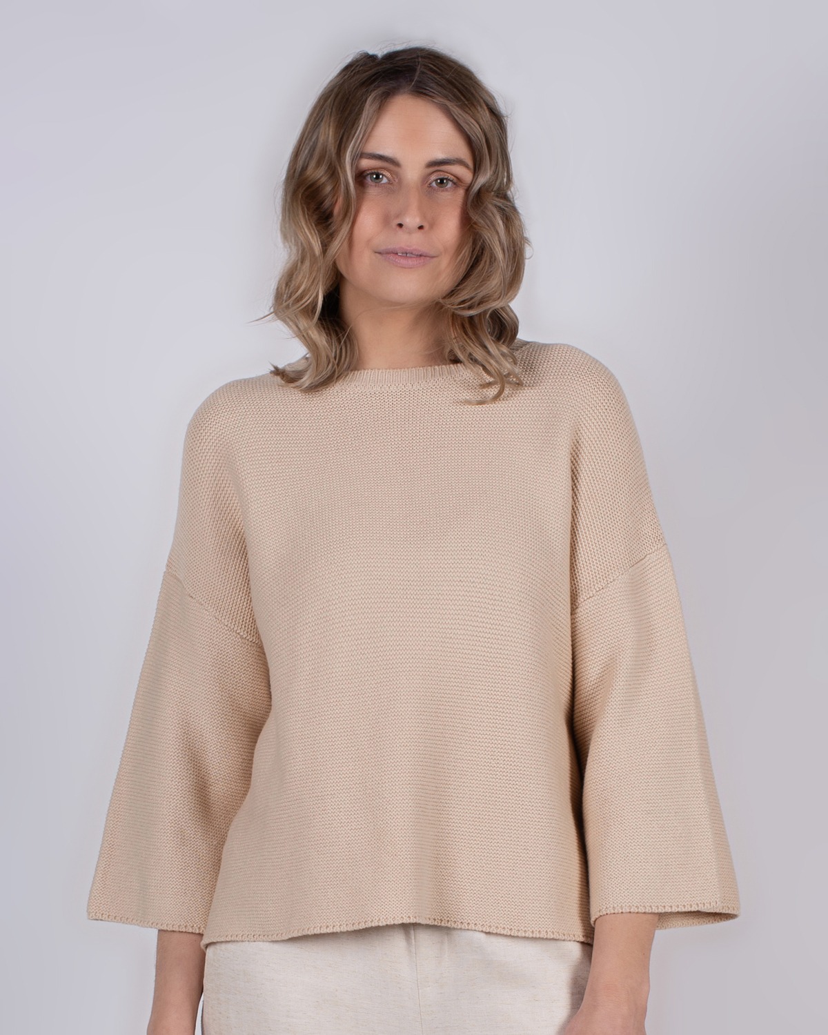marlien_studio-40 Vrouw met halflang golvend haar draagt een beige, effen trui met wijde mouwen tegen een neutrale achtergrond.