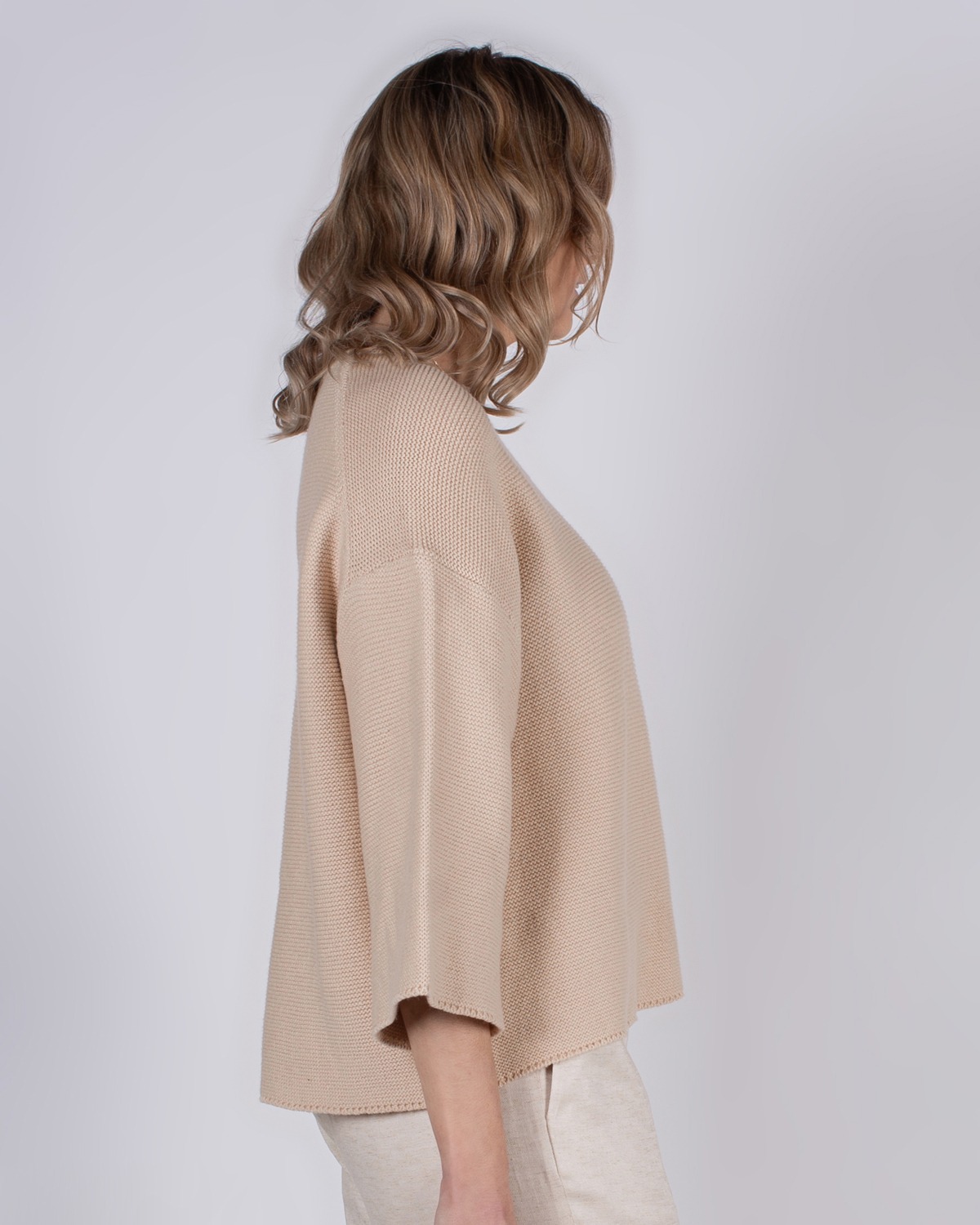 marlien_studio-41 Een vrouw met lichtbruin haar draagt een beige trui en staat zijwaarts tegen een grijze achtergrond.