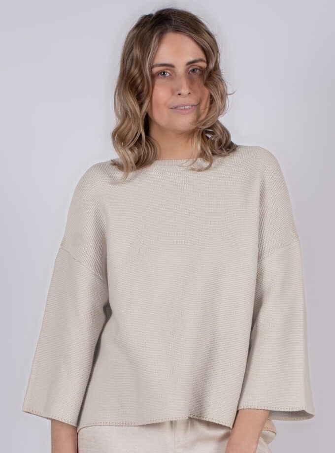 Vrouw poseert in een offwhite, ruimvallende trui met wijde mouwen tegen een neutrale achtergrond.