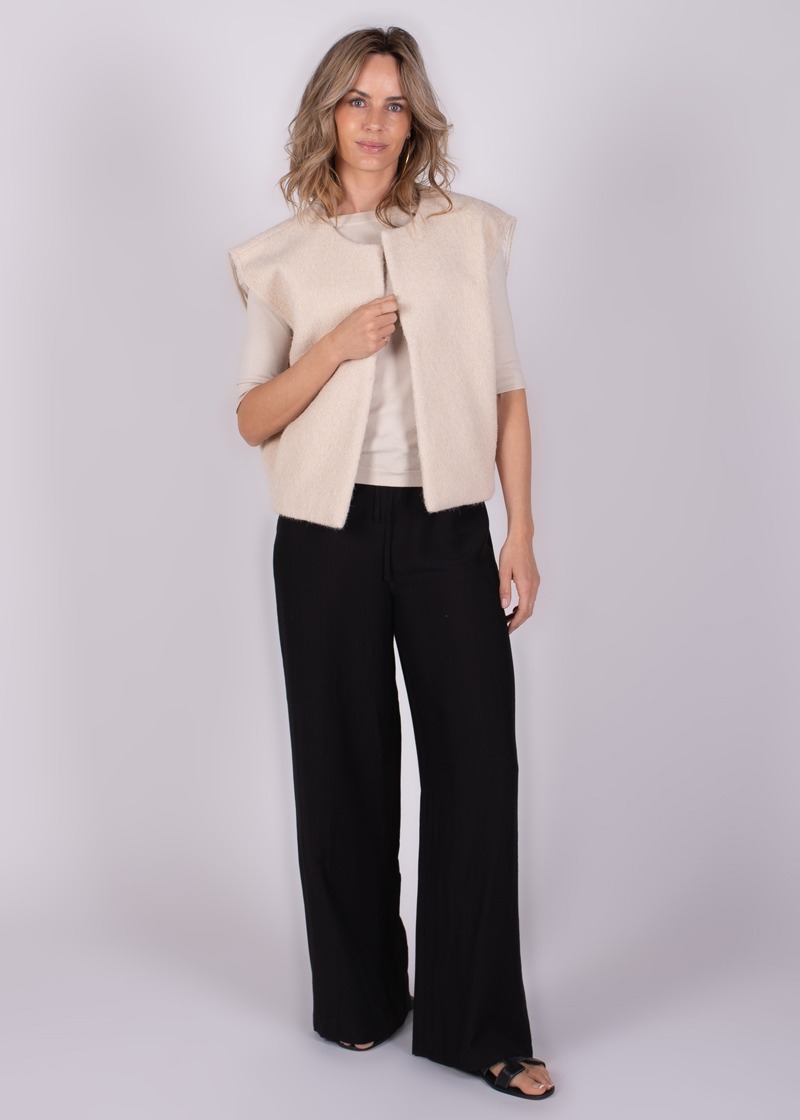 Een vrouw draagt een beige vest over een lichte top en zwarte broek, gefotografeerd in een studio tegen een grijze achtergrond.
