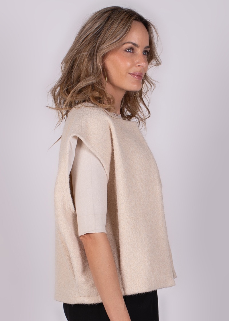 Vrouwelijk model in zijaanzicht poseert in een beige, mouwloze top met een lichte achtergrond.