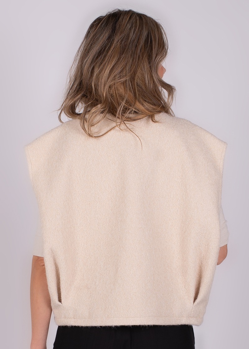 Achteraanzicht van een persoon met halflang haar, gekleed in een mouwloze, beige wollen top tegen een neutrale achtergrond.