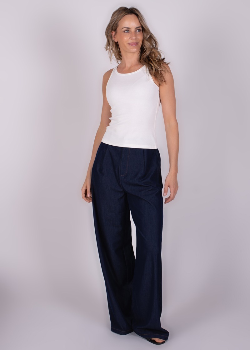 Model poseert in een witte tanktop en donkerblauwe, wijde broek tegen een neutrale achtergrond.