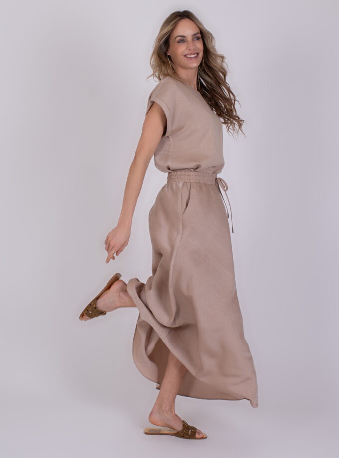 Vrouw poseert glimlachend in een beige Aba Pale 2 outfit met een lange rok en sandalen.