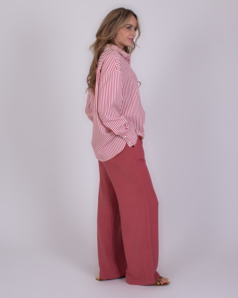 Almeria old pink 4 Vrouw in een rood-wit gestreeerde blouse en wijde, oudroze Almeria-broek poseert zijwaarts tegen een neutrale achtergrond.