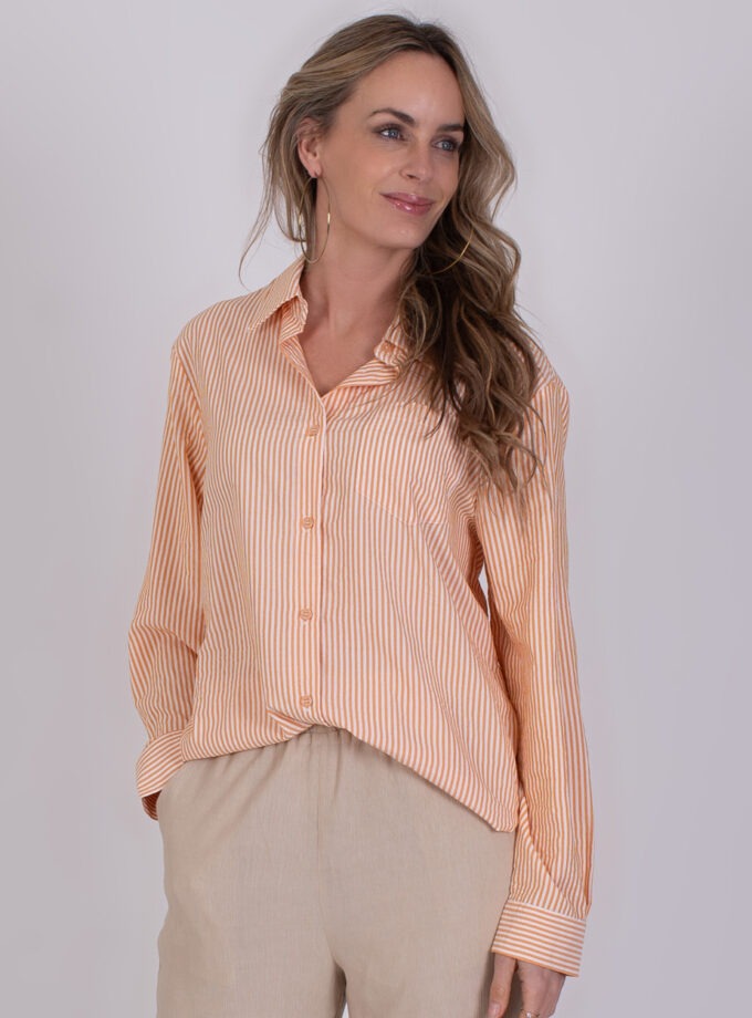 Vrouw draagt een oranje-wit gestreepte blouse genaamd Almeria orange en beige broek.