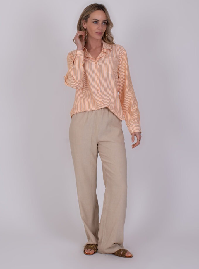 Vrouw draagt een oranje-wit gestreepte Almeria blouse met lange mouwen en beige linnen broek.