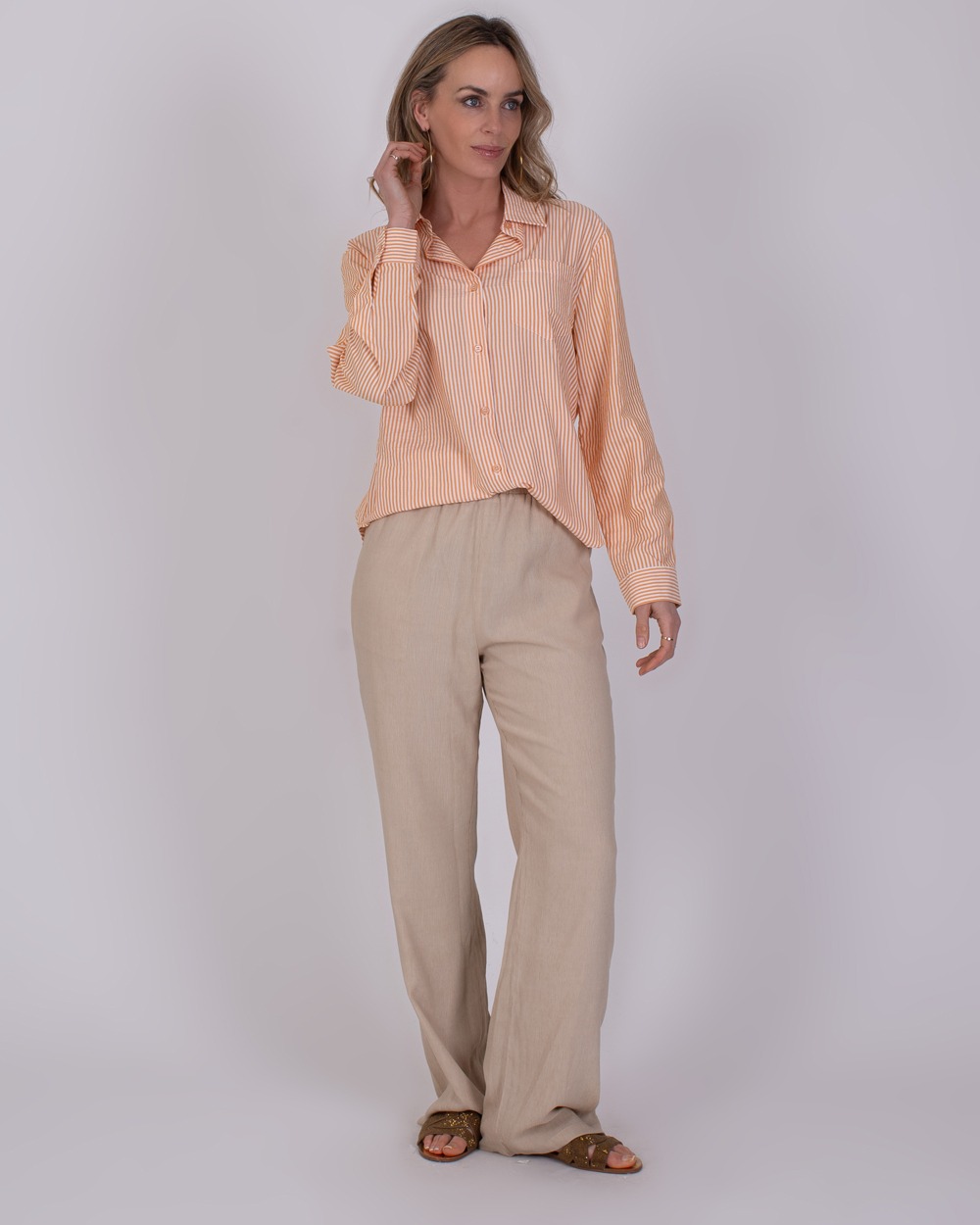 Almeria orange 2 Vrouw draagt een oranje-wit gestreepte Almeria blouse met lange mouwen en beige linnen broek.