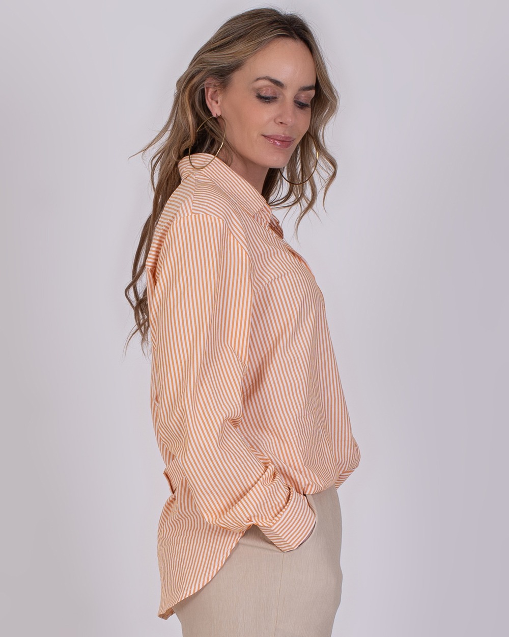 Almeria orange 3 Vrouw draagt een losvallende, oranje-wit gestreepte blouse genaamd Almeria Orange 3 met beige broek.