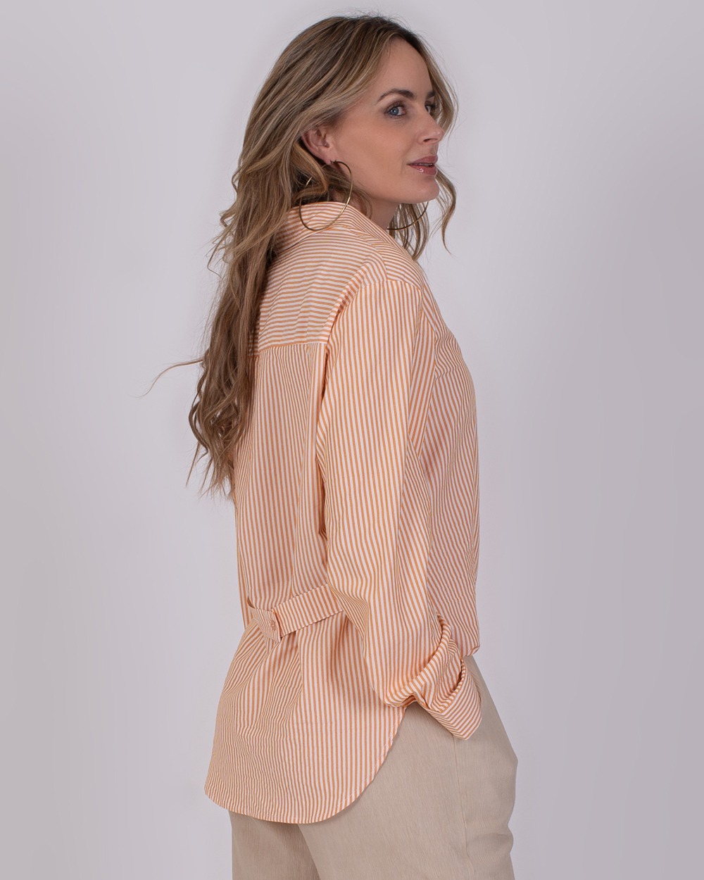 Almeria orange 4 Vrouw draagt een oranje-wit gestreepte blouse genaamd Almeria orange, gecombineerd met een beige broek.