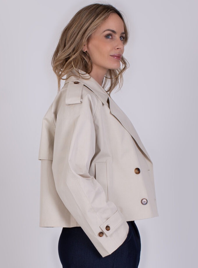 Vrouw draagt een korte beige trenchcoat genaamd Aosta Pale 3 met dubbele knopenrij en epauletten.