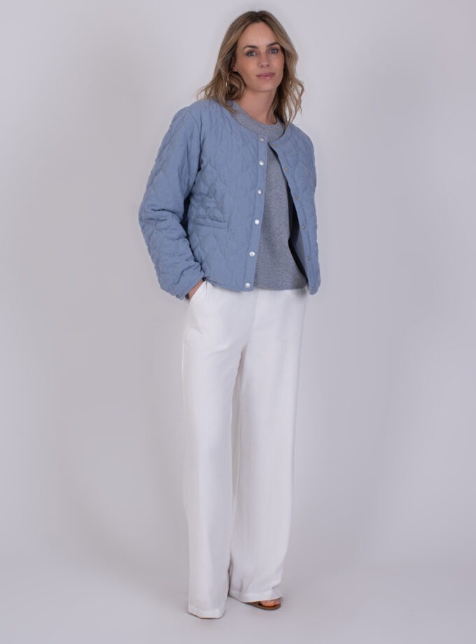 Vrouw draagt een lichtblauwe gewatteerde 'Burgas soft blue' jas, grijze top en witte wijde broek.
