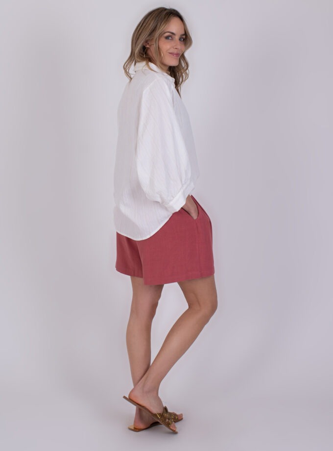Vrouw draagt een witte Busan off white blouse met roze shorts en slippers tegen een neutrale achtergrond.