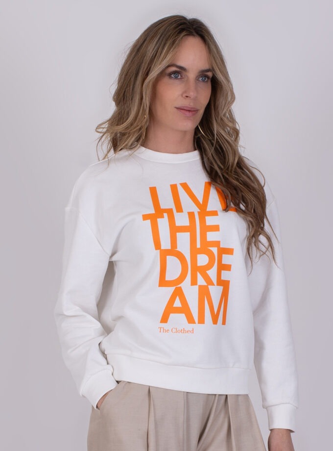Vrouw draagt een offwhite Capri-trui met feloranje tekst "LIVE THE DREAM" op de borst.
