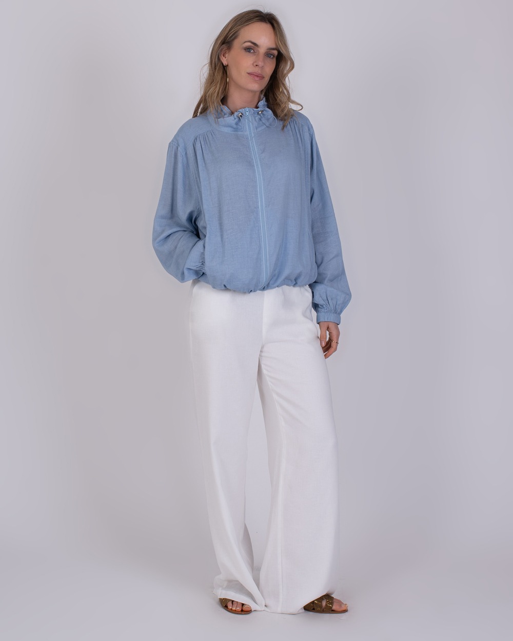 Edessa soft blue 1 Vrouw draagt een lichtblauwe Edessa-blouse met ritssluiting en witte, wijde broek.