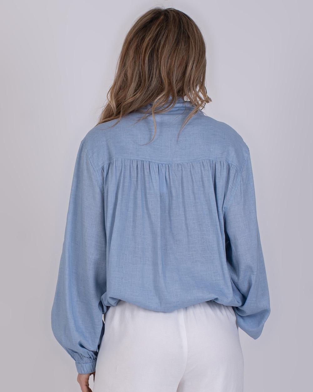 Edessa soft blue 3 Achteraanzicht van een vrouw die een lichtblauwe Edessa-blouse en witte broek draagt.