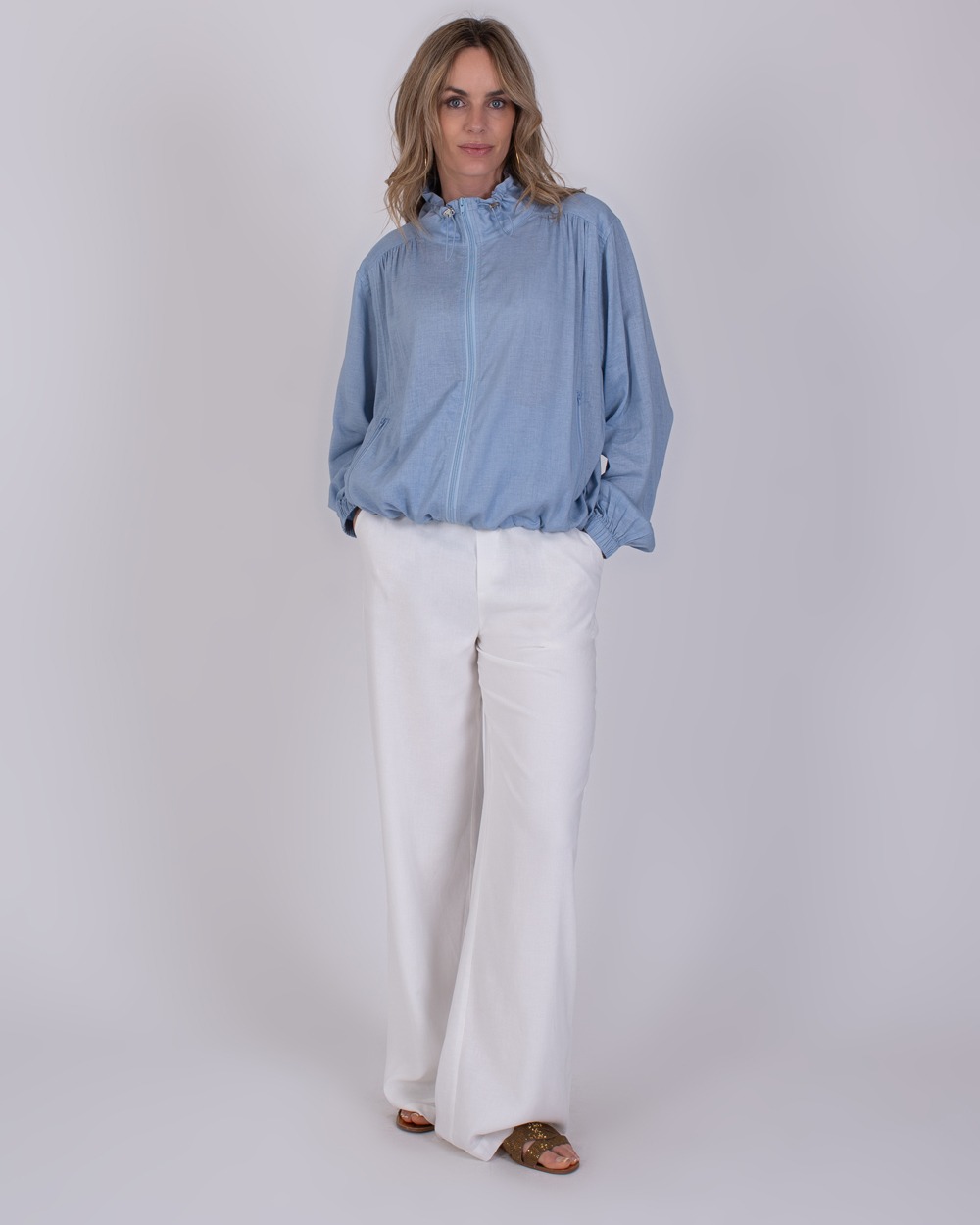 Edessa soft blue 4 Vrouw draagt een lichtblauwe Edessa soft blue blouse met witte, wijde broek en sandalen.