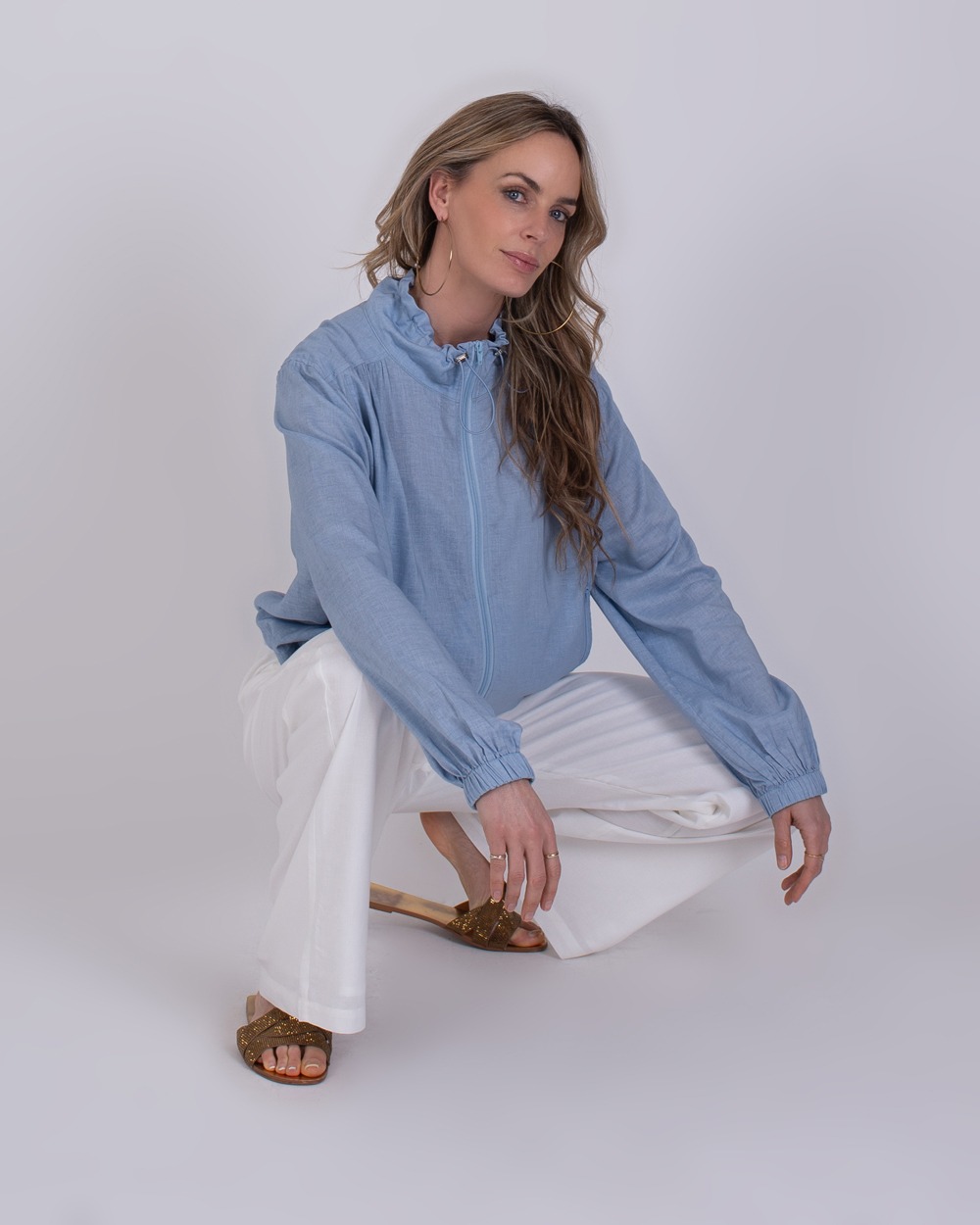 Edessa soft blue 5 Vrouw draagt een lichtblauwe Edessa soft blue blouse met ruches en witte broek terwijl ze gehurkt poseert.